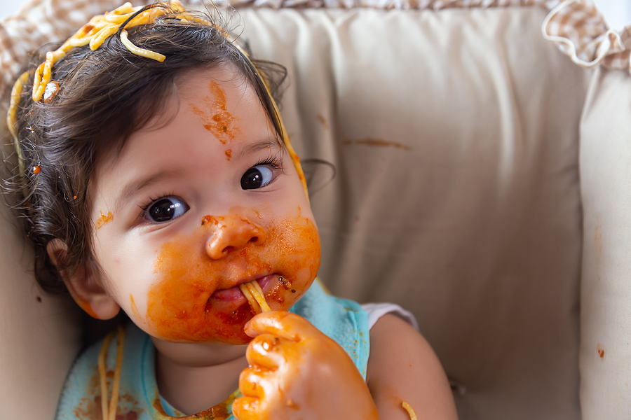 Adorable little toddler girl or infant baby eating delicious spaghetti food with tomato sauce on baby chair after he opt out washing his face, Cute infant girl practice eating food . Mix race daughter look dirty hand